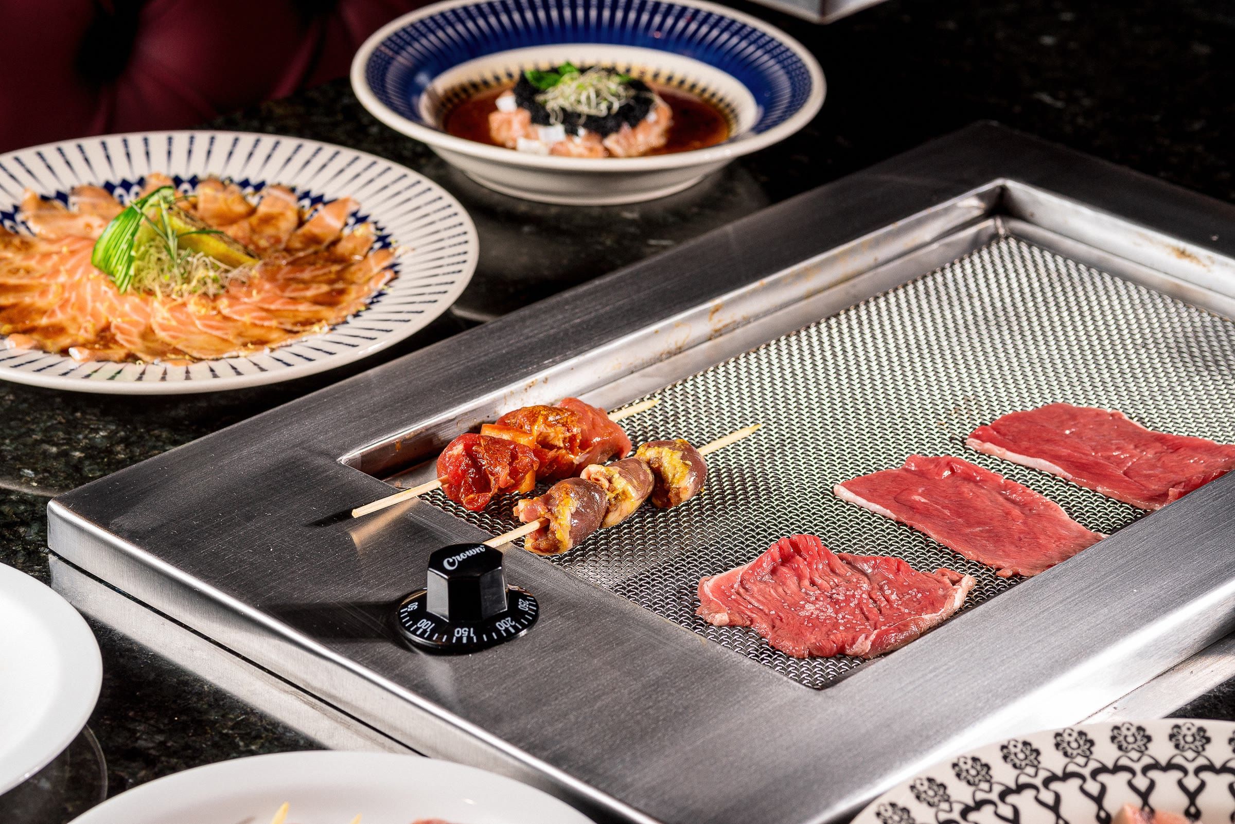 Chapa de inox com espetinho e duas fatias de carne crua, pratos decorados ao redor