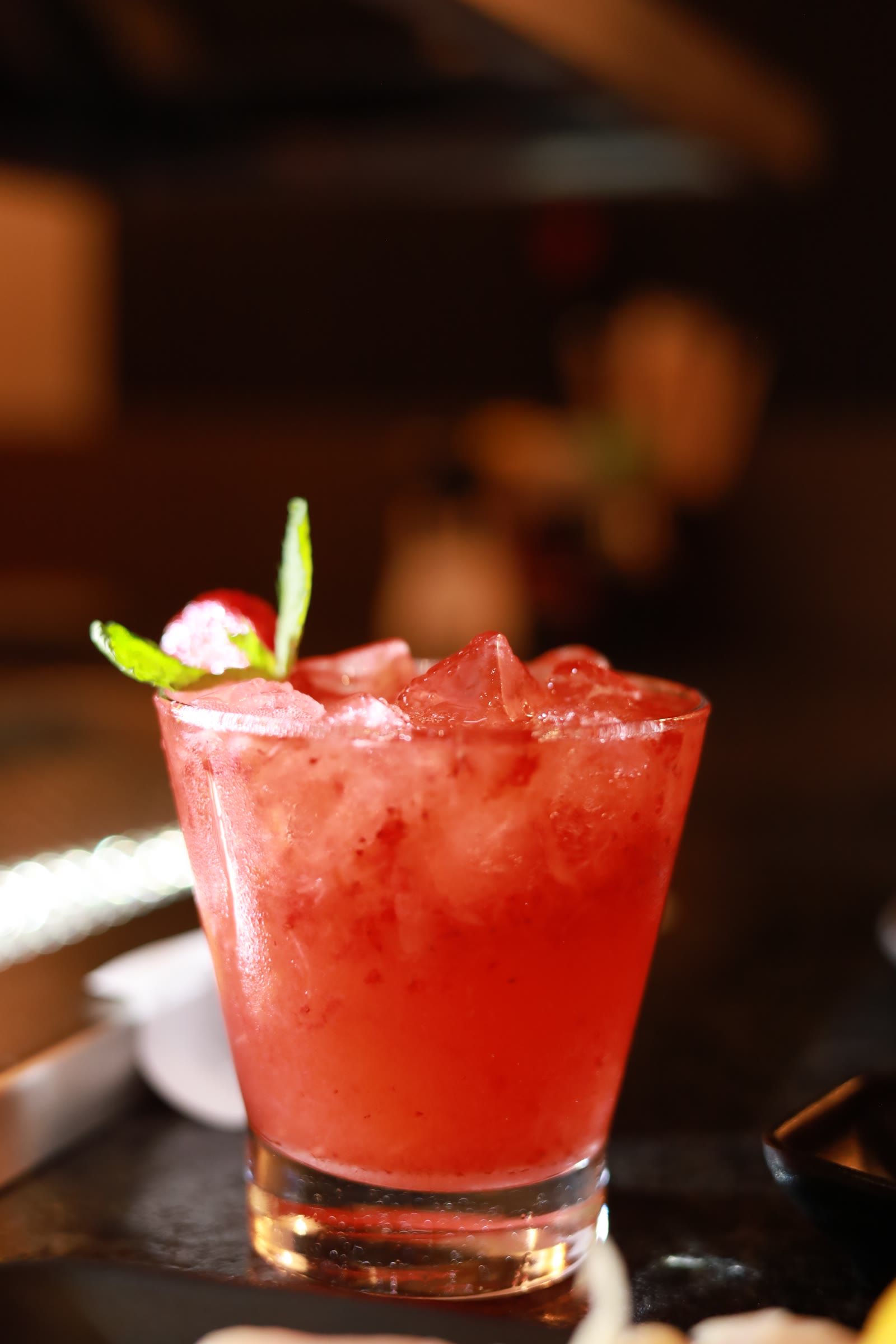 Drink vermelho com gelo triturado, morango e hortela em copo baixo, fundo escuro desfocado
