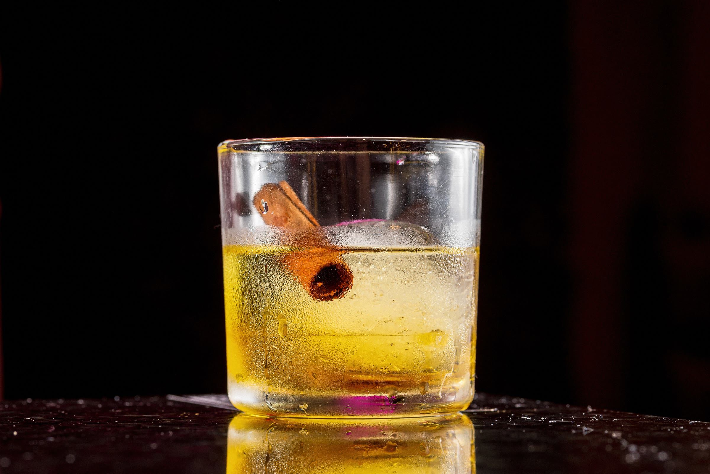 Copo de whisky ambar com canela e gelo sobre fundo preto, reflexo na mesa polida