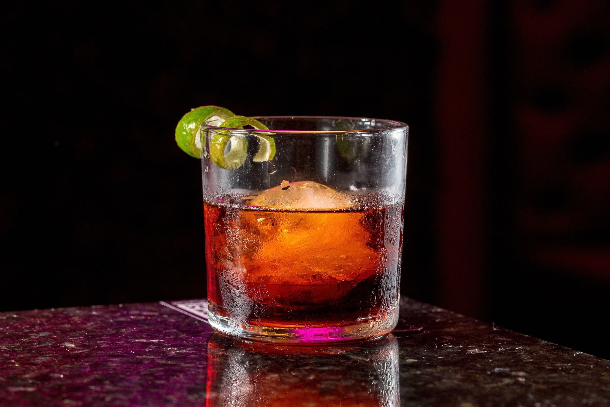 Coquetel vermelho tipo negroni com casca de limao e gelo esferico, fundo escuro e reflexo