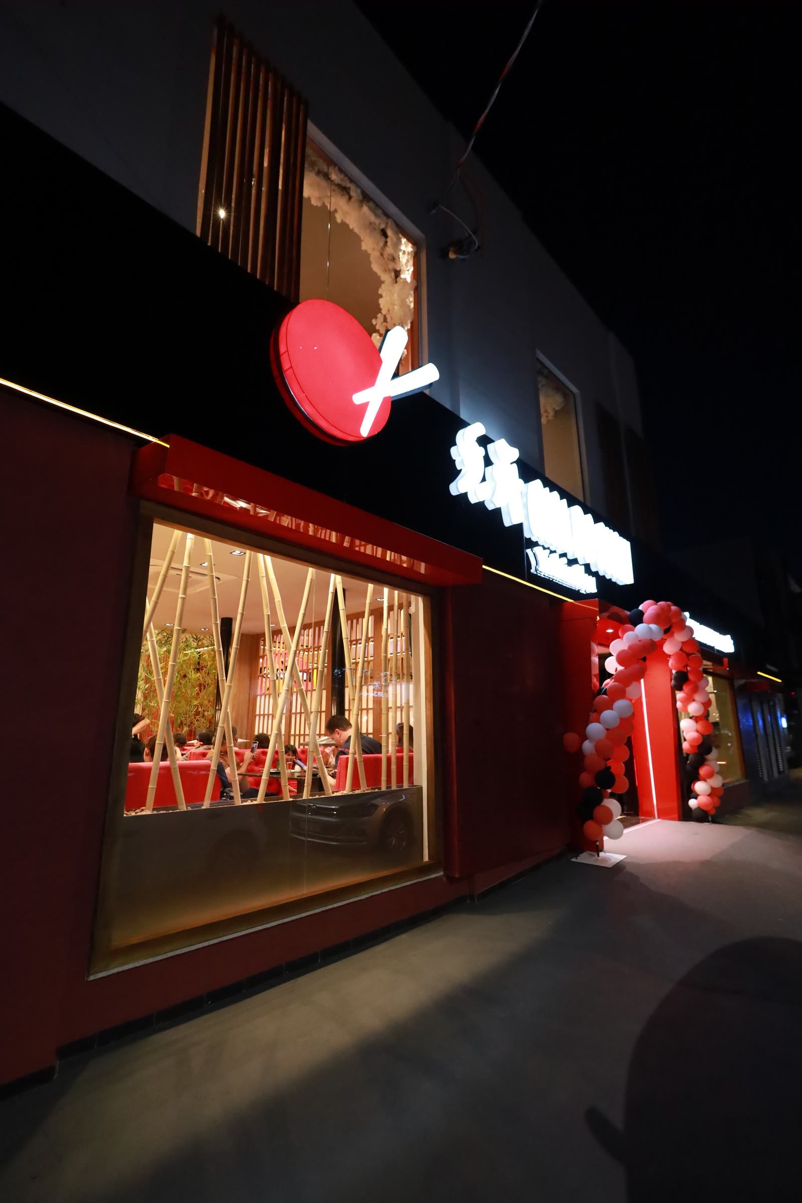 Fachada noturna em ângulo, logo vermelho aceso no painel preto e vitrine iluminada