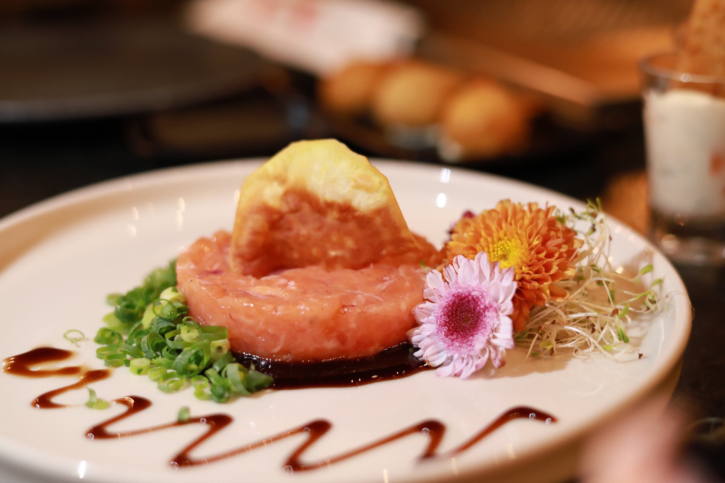 Tartar de salmao com flor comestivel e brotos em prato branco, bolinhos e copo desfocados ao fundo