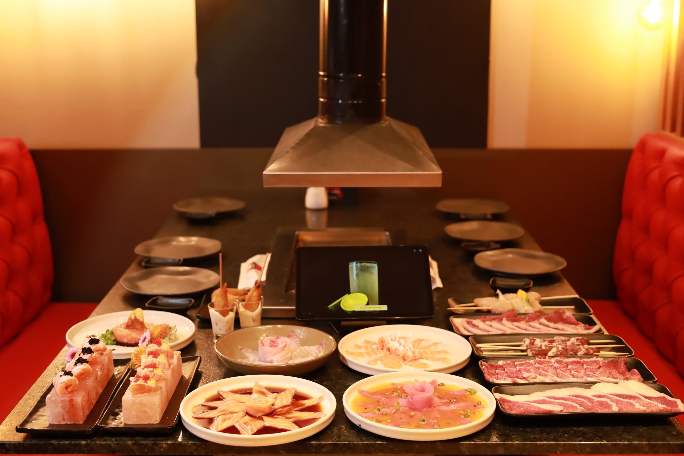 Mesa de rodízio posta com bandejas de carne crua fatiada, pratos de sashimi e acompanhamentos sob a coifa, entre bancos capitonê vermelhos