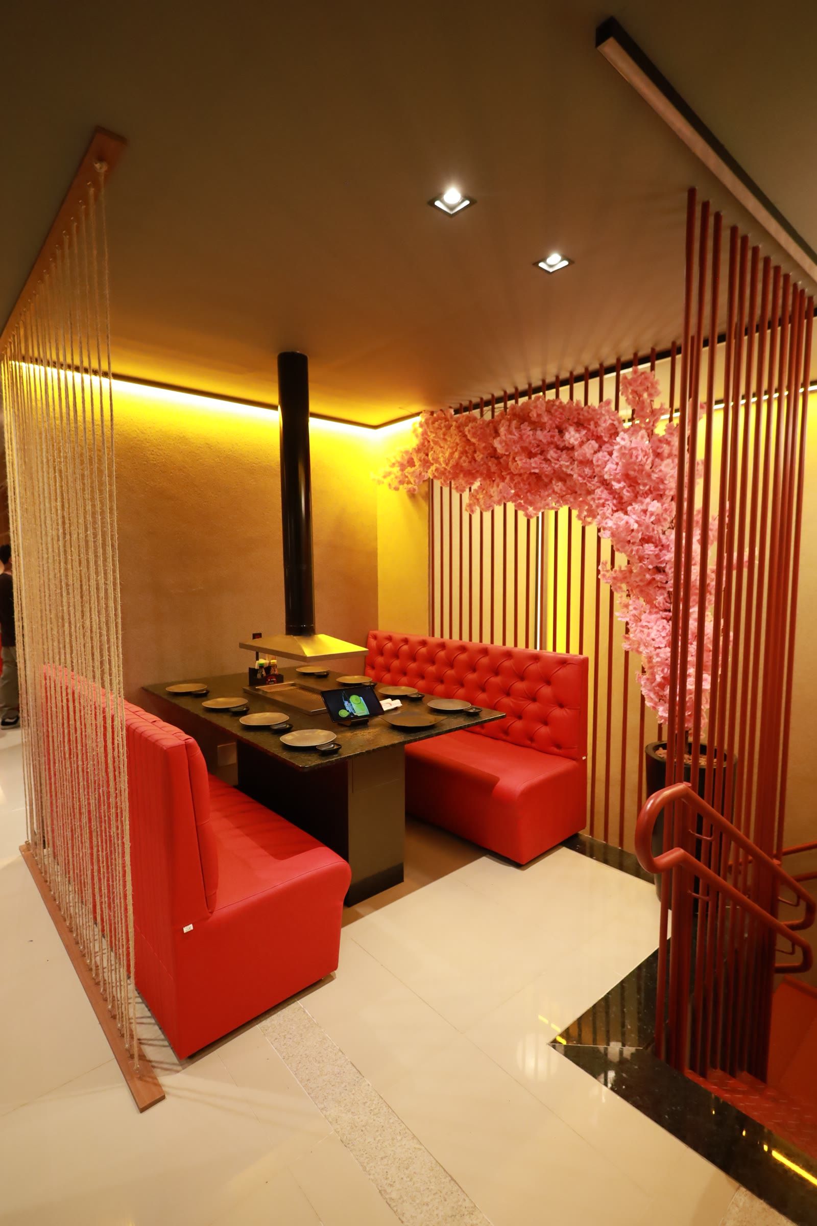 Mesa de yakiniku com sofas vermelhos capitone, cerejeira rosa e biombo de ripas vermelhas ao fundo