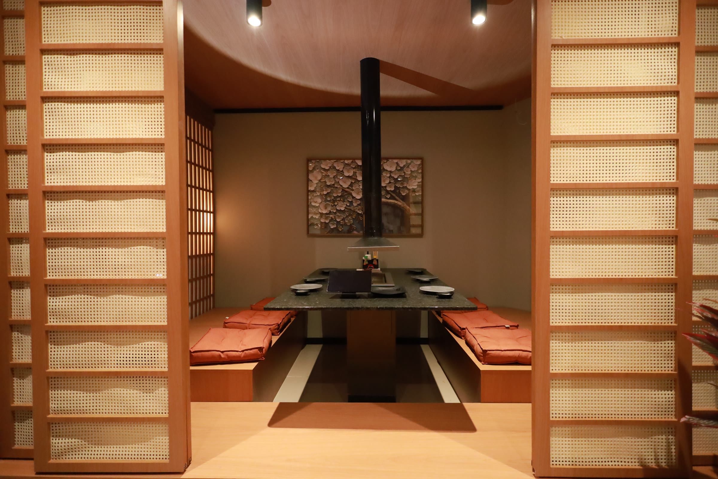 Sala privativa em tatame vista entre portas shoji, mesa rebaixada com grelha e almofadas laranja