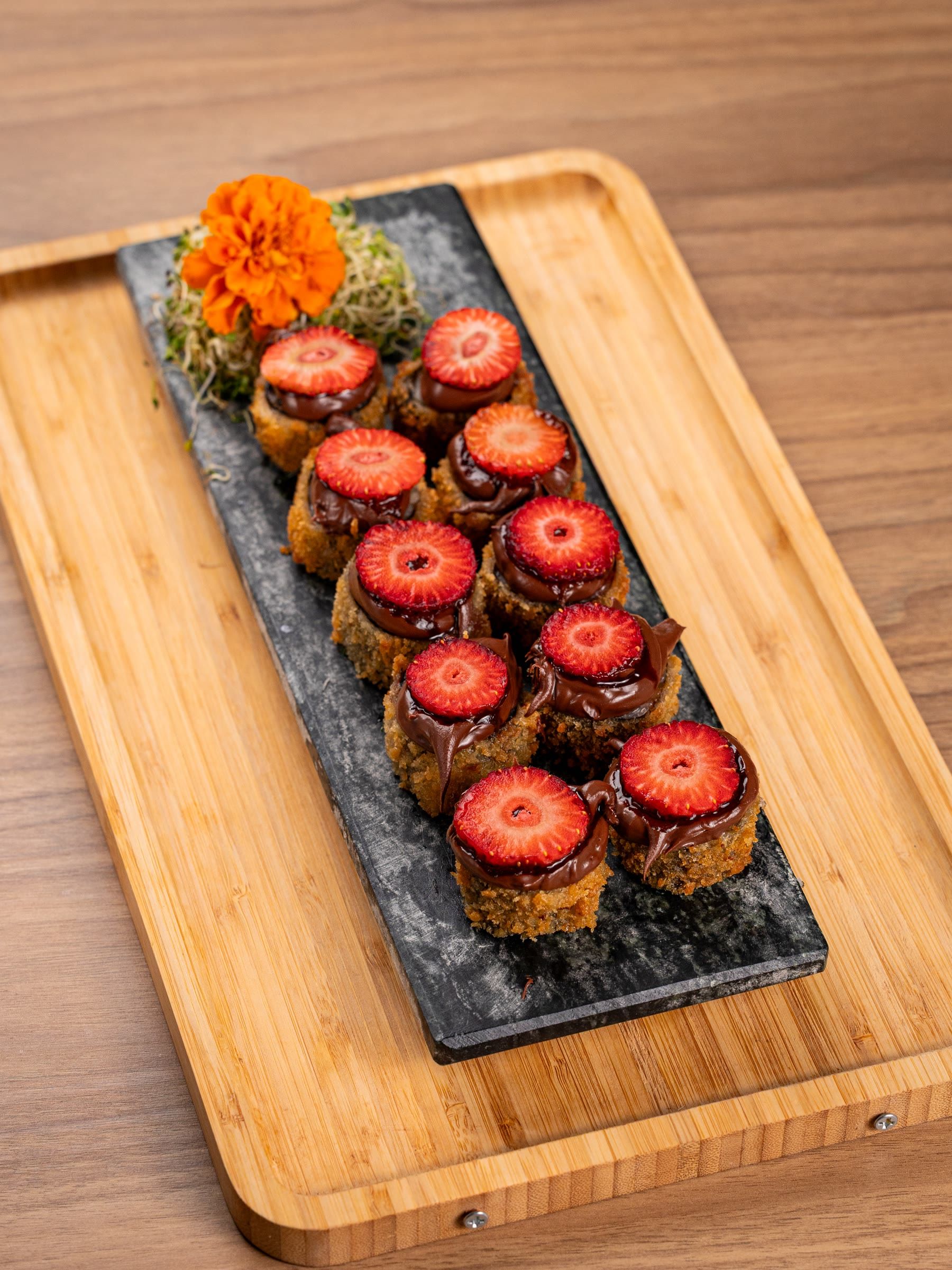 Fileira de rolls doces com calda de chocolate e morangos fatiados em ardosia sobre tabua de bambu