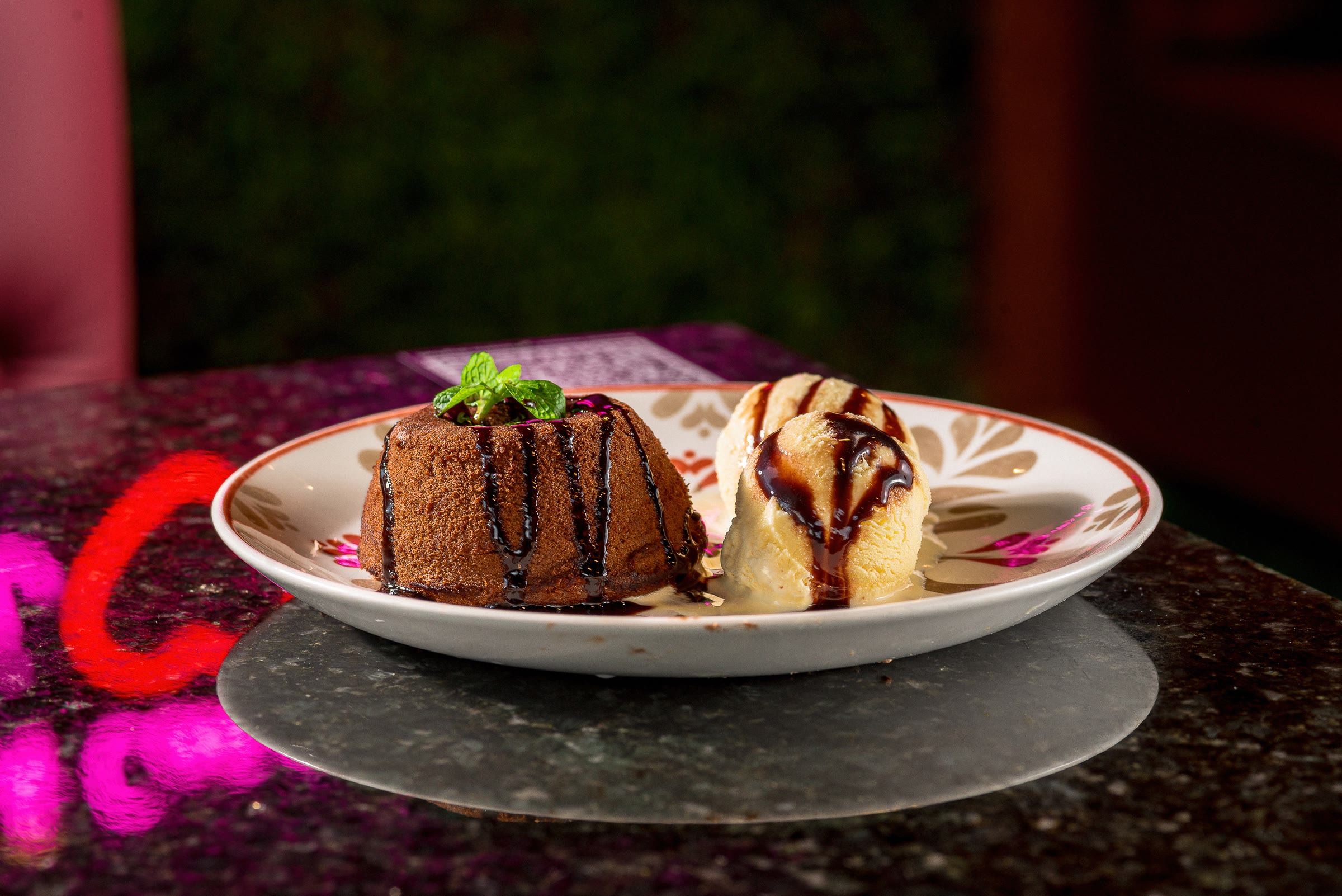 Bolo de chocolate com calda e sorvete em prato branco floral, fundo escuro com neon rosa