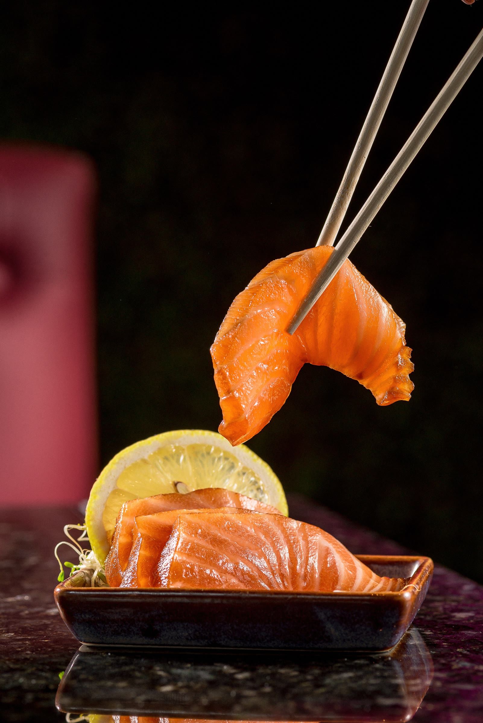 Hashi de metal ergue fatia brilhante de salmão acima da barquinha de sashimi, fundo preto