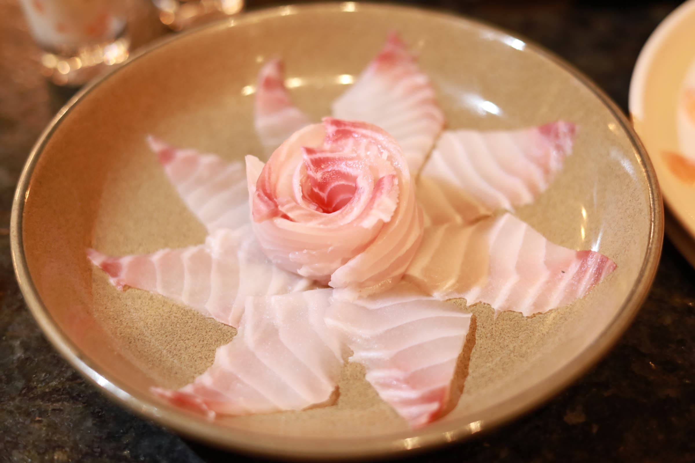 Fatias finas de peixe branco arranjadas em forma de flor rosada sobre prato dourado