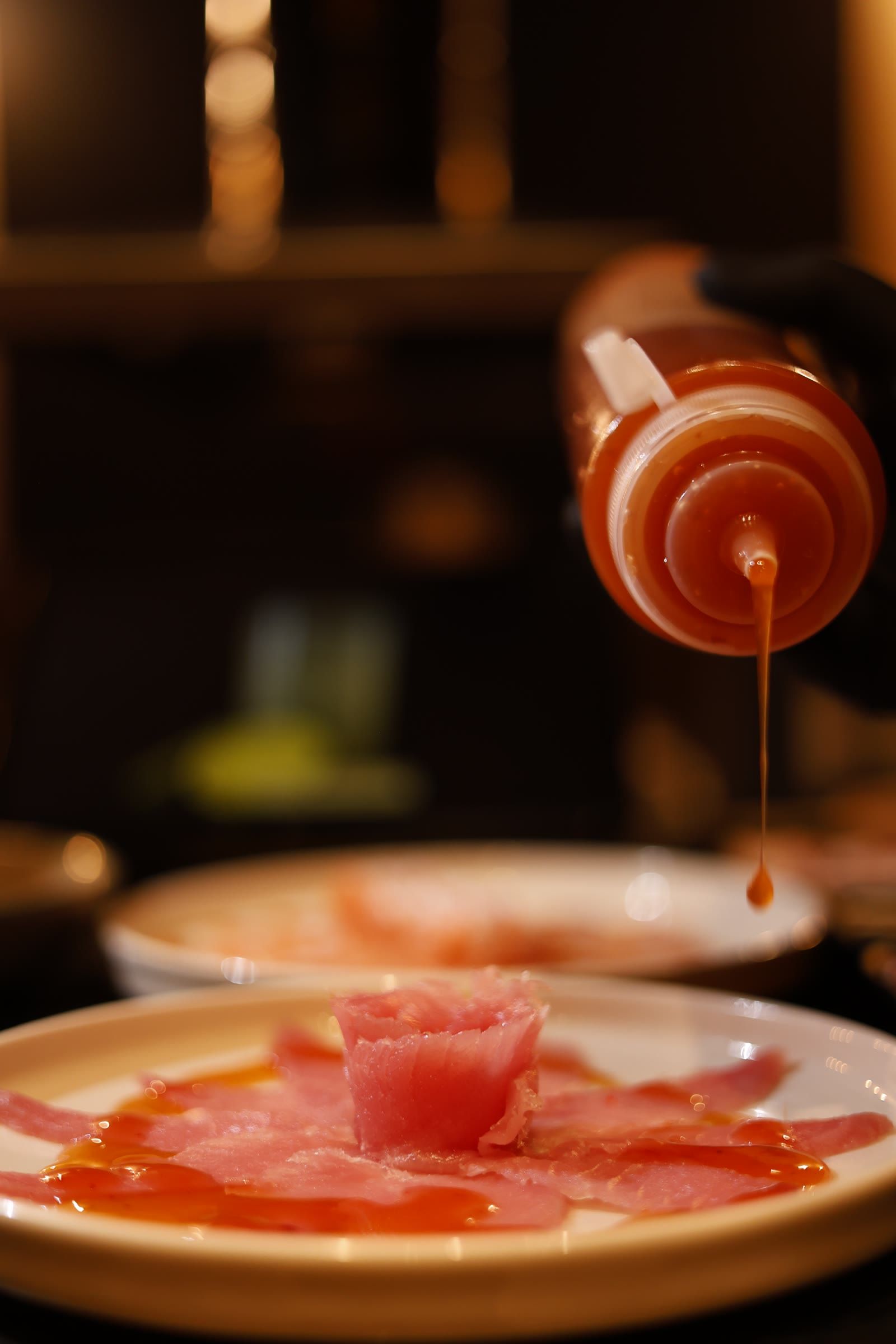 Mão enluvada despeja fio de molho alaranjado sobre carpaccio de peixe com rosa ao centro.