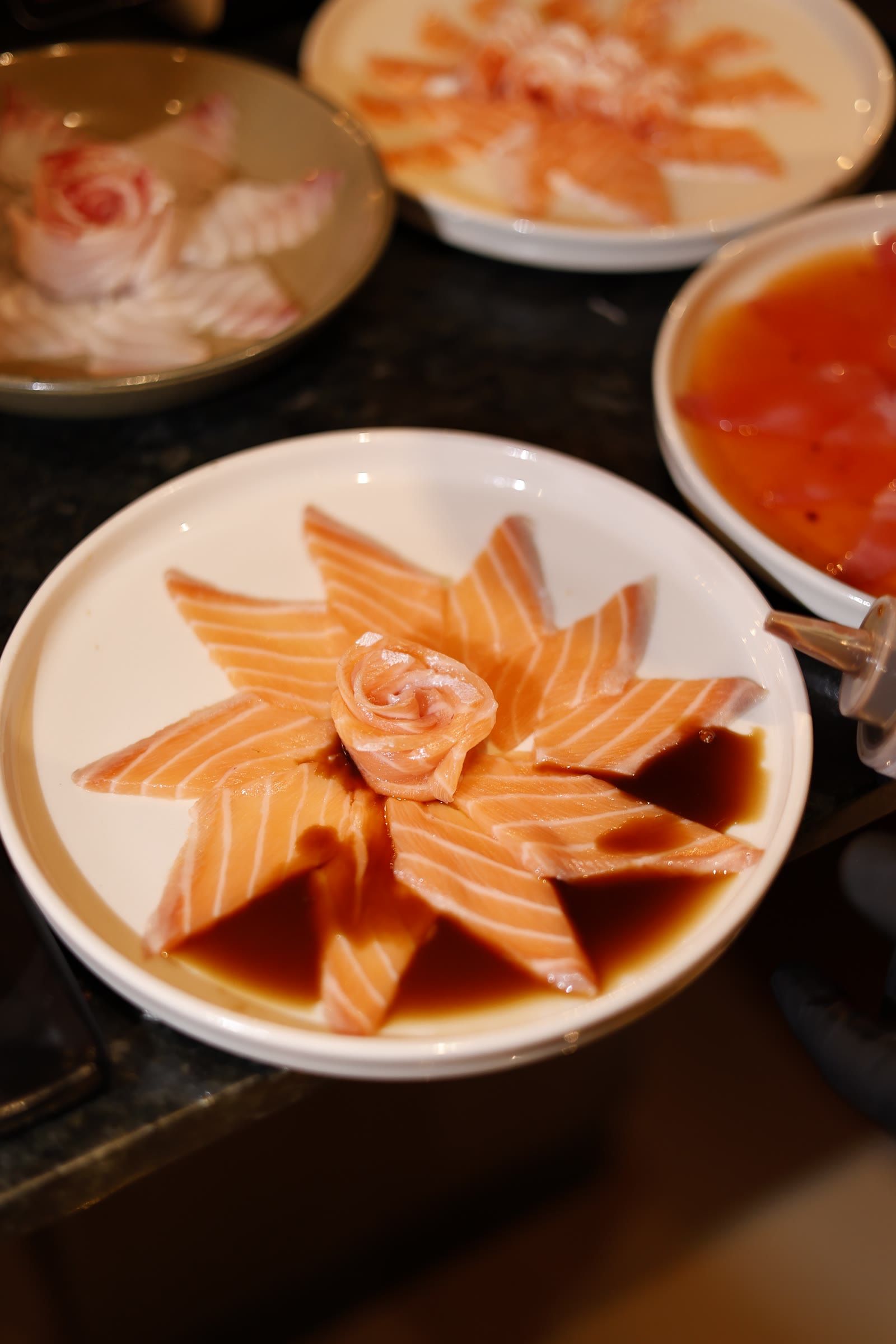 Prato de sashimi de salmão em flor sobre shoyu, cercado por outros pratos no balcão.