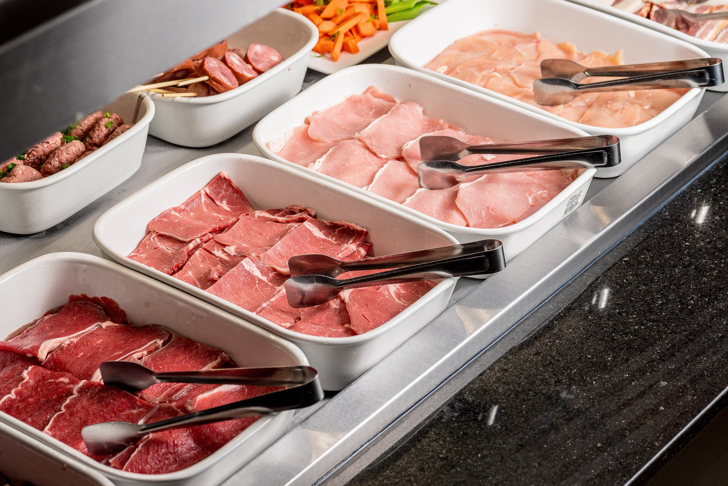 Bandejas brancas com carne crua fatiada e pegadores pretos ao lado de cubas de legumes coloridos