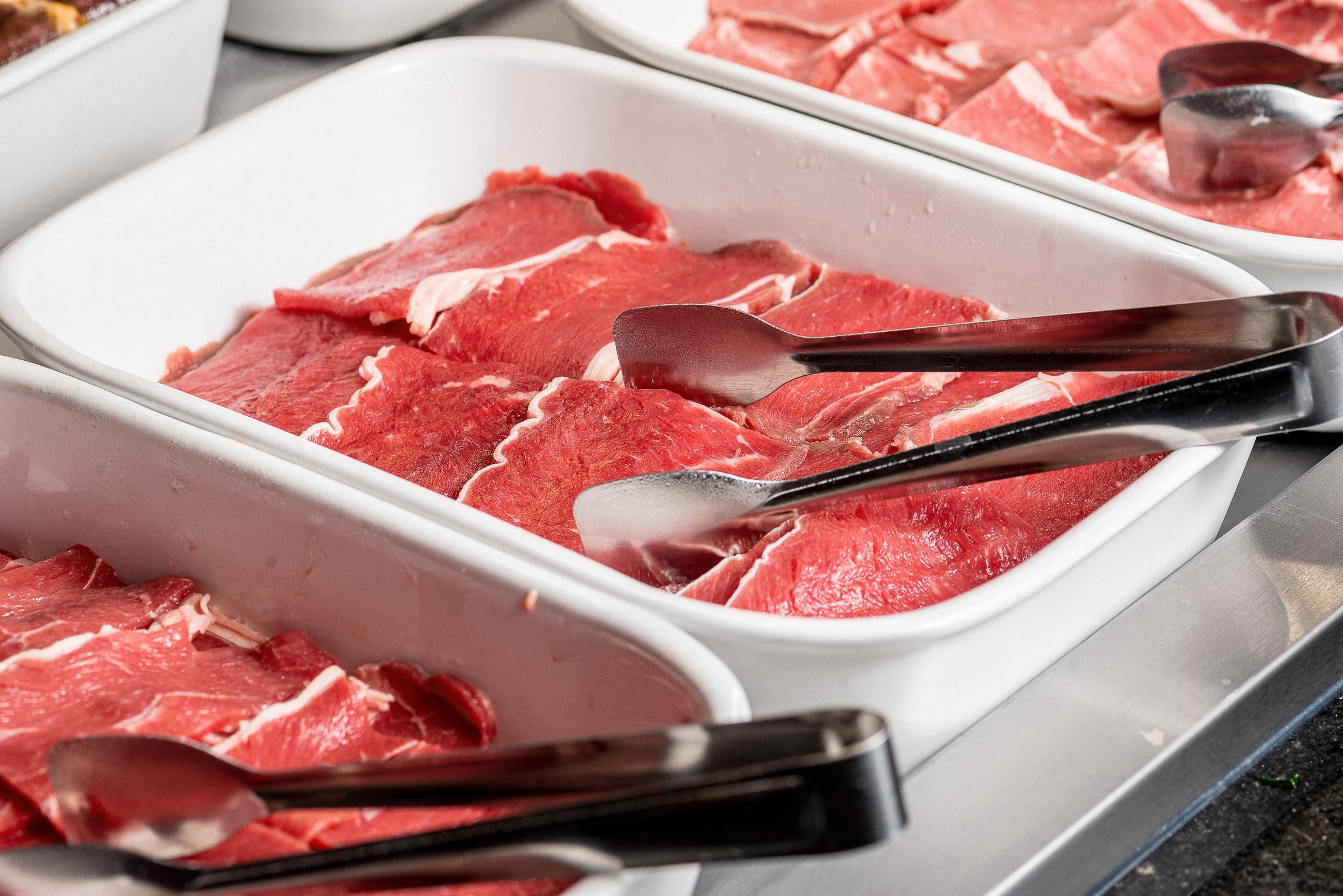 Close da bandeja com fatias finas de carne crua marmorizada e pegador preto no balcao refrigerado