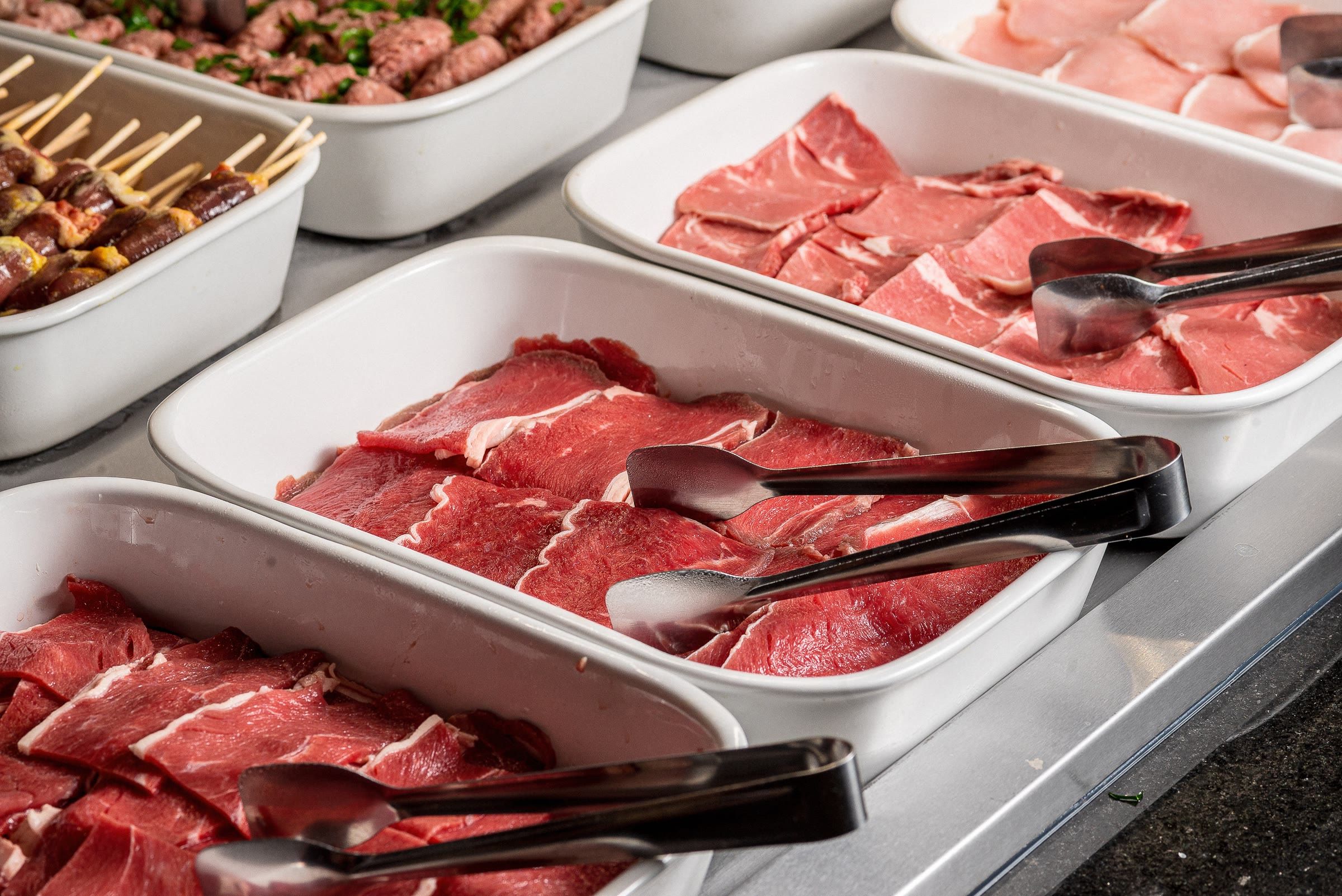 Bandejas de carne crua fatiada com pegadores e espetinhos temperados na lateral do balcao de grelhar