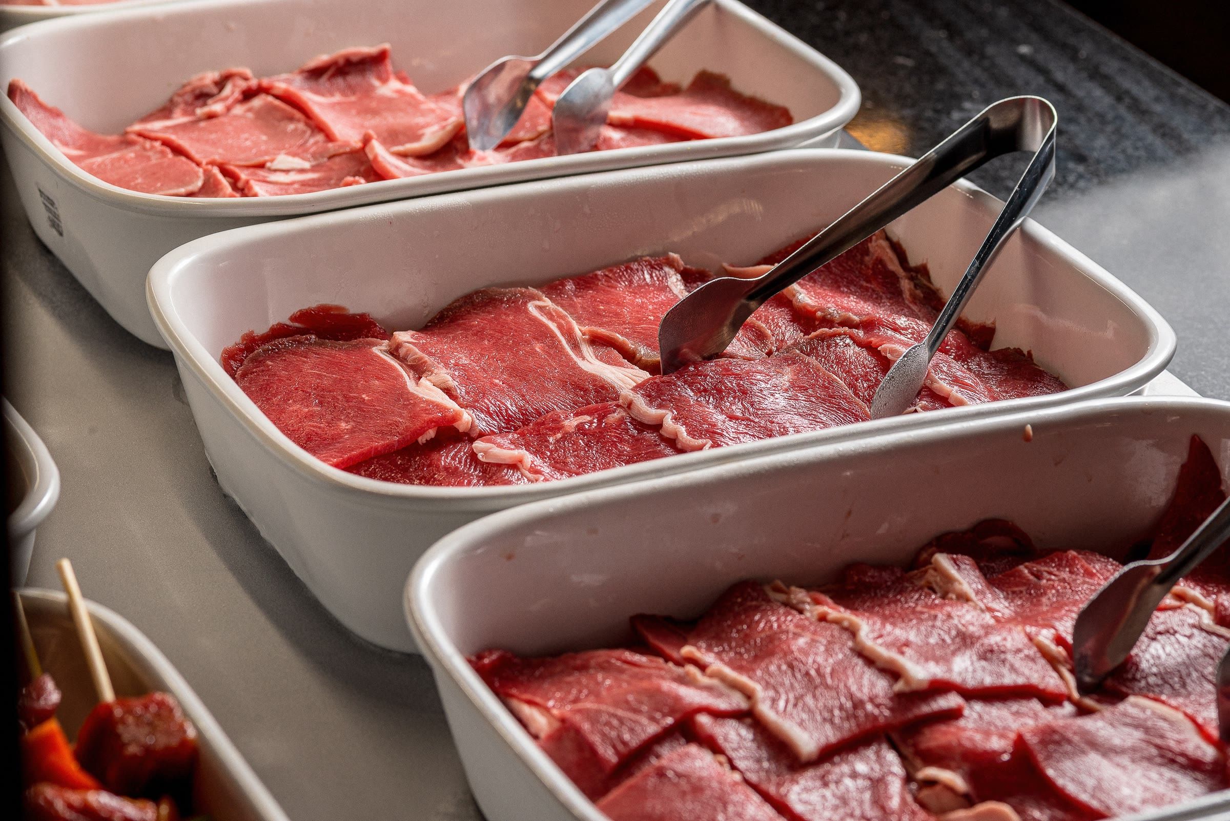 Cubas brancas com fatias finas de carne bovina crua vermelha e pegadores de inox no balcao
