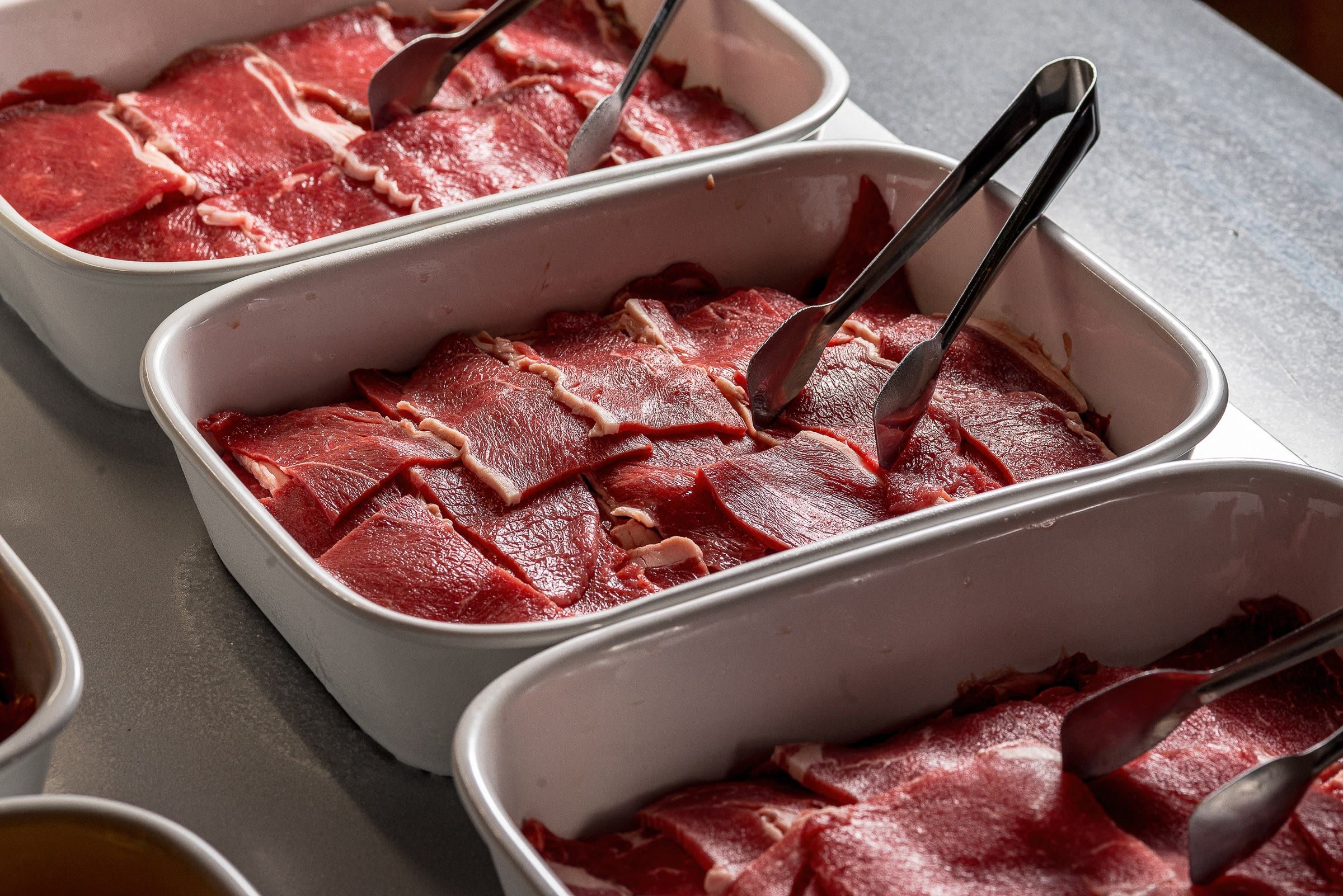 Tres cubas alinhadas de carne bovina fatiada crua com pegadores de inox, luz quente sobre o balcao